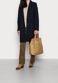 Frau in navy Blauem Mantel, beigem Pullover, braunen Cordhosen und beigen Stiefeletten, hält eine beige Ledertasche vor einem schlichten Hintergrund.