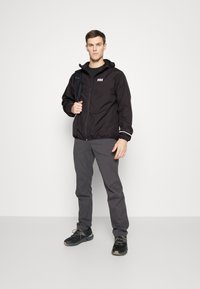 Schwarze leichte Jacke mit Kapuze, weißen Akzenten und Logo; kombiniert mit grauen Hosen und schwarzen Schuhen mit Schnürsenkeln und strukturiertem Sohlenprofil.
