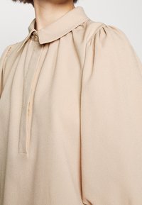 Beige blus med krage, knappstängning och volangärmar. Tillverkad av mjukt, lätt material med en slät textur.