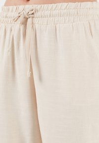 Soft beige fabric joggers with an elastic waistband and tie detail. Features a textured surface with subtle horizontal lines.