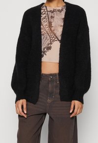 Černý, huňatý kardigan s otevřeným předním dílem a dlouhými rukávy. Nositý přes vzorovaný crop top, zkombinovaný s tmavě hnědými kalhotami s vysokým pasem.