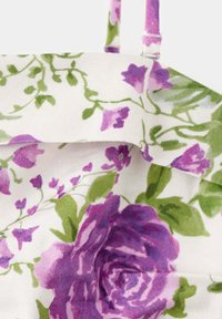 Gros plan d’un tissu avec motif floral de roses violettes et feuilles vertes sur fond blanc, montrant une partie d’une sangle attachée par un anneau en métal.