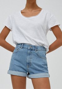 Person med en enkel vit t-shirt med kort ärm instoppad i high-waisted ljusblå denimshorts med upprullad fåll framför en neutral bakgrund.