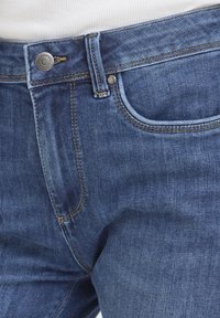 Gros plan sur la taille d'un jean en denim bleu avec un bouton argenté, une poche avant et une passants de ceinture, associé à un haut blanc côtelé.