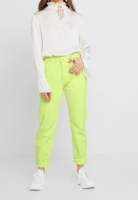 Blusa blanca con un escote volante y detalle de ojal, combinada con pantalones verdes neón brillantes y zapatillas blancas. Texturas suaves en todo.