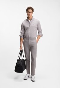 Ανοιχτό γκρι πουκάμισο με κουμπιά και μαύρο λογότυπο, σε συνδυασμό με ανοιχτό γκρι παντελόνια. Το μοντέλο κρατάει μια μαύρη τσάντα και φοράει λευκά αθλητικά παπούτσια με γκρι λεπτομέρειες.