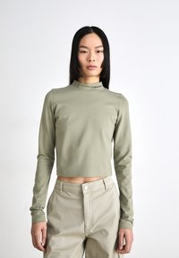 Long-sleeve, cropped top in olive green, made of smooth fabric; high neckline with black "Calvin Klein" logo on the collar.