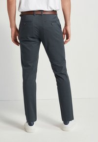Pantaloni grigi scuri con una vestibilità Slim, dotati di due tasche posteriori con chiusura a bottone. Il tessuto presenta una sottile texture. Indossati con sneakers bianche.