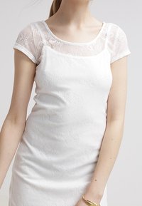 Bílé upnuté šaty s texturovanou tkaninou a průhlednými krajkovými krátkými rukávy. Mají kulatý výstřih a hladký, jednuduchý design.