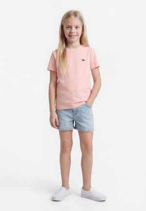 Jong meisje staand, gekleed in een roze shirt met korte mouwen, licht denim korte broek en witte instapschoenen, glimlachend met één hand in de zak.