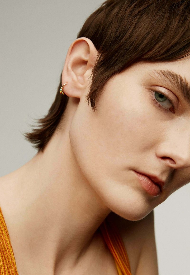 Pendiente de aro dorado con un pequeño acento de piedra multicolor, llevado en una oreja. El primer plano muestra piel suave y cabello corto y oscuro.