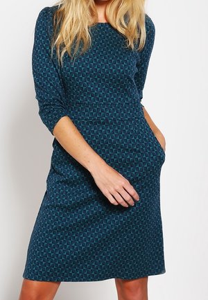 Femme portant une robe couleur sarcelle jusqu'aux genoux avec des manches trois-quarts et un motif géométrique, une main dans une poche et l'autre reposant sur la jupe.