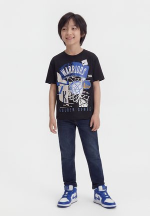 Garçon souriant, portant un t-shirt noir des Golden State Warriors, un jean foncé et des baskets montantes bleu et blanc, debout sur un fond blanc.