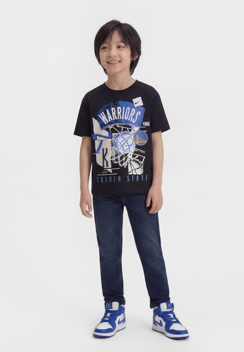 Αγόρι που χαμογελά, φοράει μαύρο μπλουζάκι Golden State Warriors, σκούρα τζιν και μπλε-λευκά αθλητικά παπούτσια με ψηλό πέλμα, στέκεται πάνω σε λευκό φόντο.