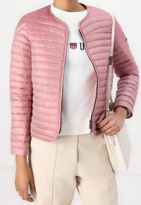 Ljusrosa isolerad jacka med dragkedja, vågrätt sömnad och rund halsringning. Har en svart dragkedja och en slät textur.