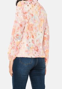 Kvinna som bär en genomskinlig blus med lång ärm i pastellfärgade blommor och guldprickigt mönster, ihop med mörkblå denimjeans, visad bakifrån.