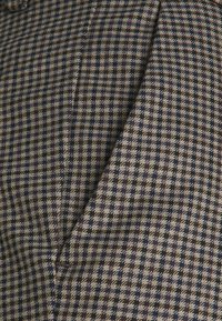 Tissu en laine à motif pied-de-poule, présentant des carrés alternés bleu foncé, marron et crème. Texture lisse, sensation douce.