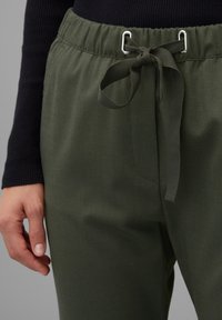 Hand som vilar bredvid olivgröna byxor med elastiskt midjeband och knytband av tyg mot en grå bakgrund.