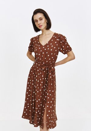 Eine Frau posiert selbstbewusst in einem braunen Kleid mit Punkten, das einen V-Ausschnitt und eine gebundene Taille hat, vor einem schlichten weißen Hintergrund.