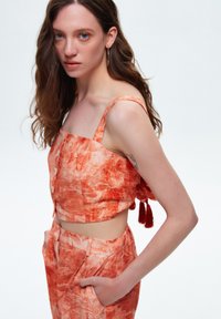 Oranje cropped top met knopen en verstelbare bandjes, gecombineerd met bijpassende broek. De stof heeft een bloempatroon en textuur.