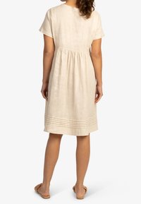 Beige lněné šaty s krátkými rukávy, elastickým pasem a plisovaným lemem. Jednoduchý, volný design pro pohodlí. Nosené s plochými sandály.