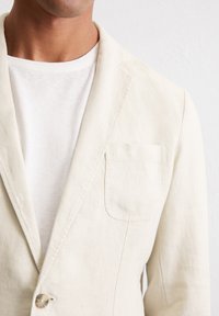 Beige Leinenblazer mit einer einzelnen Brusttasche, mit Revers und einer Zwei-Knopf-Verschluss, getragen über einem weißen Rundhals-T-Shirt.