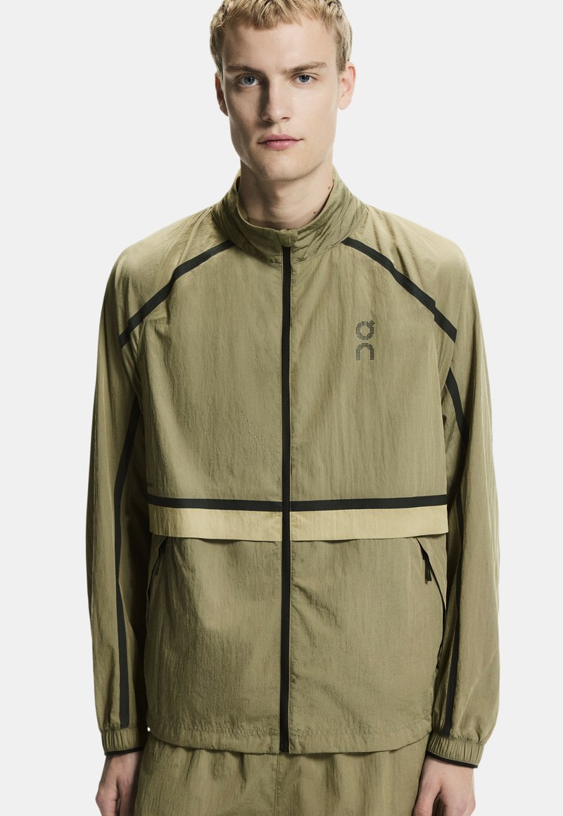 Jeune homme portant une veste vert olive clair avec une fermeture éclair noire et des détails en bande, debout devant un fond blanc uni.