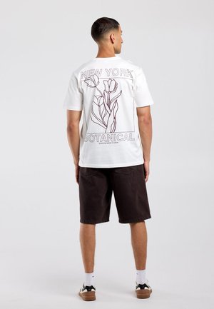 Man draagt een wit T-shirt met botanische lijntekening en tekst "NEW YORK BOTANICAL" en donkere korte broek, staat met de rug naar voren op een effen achtergrond.