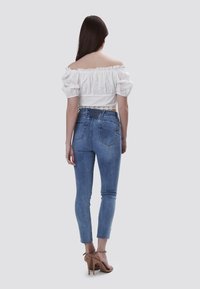 Blusa blanca de hombros al descubierto con mangas abullonadas, detalles de ojales, combinada con jeans ajustados de cintura alta en azul claro. Sandalias de tacón alto en color beige.