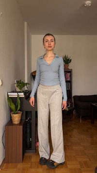 Νέα γυναίκα με ανοιχτό μπλε μπλούζα μανικιών και μπεζ παντελόνια στέκεται σε ένα σαλόνι με φυτά, βιβλία και έναν σκούρο καναπέ στο φόντο.