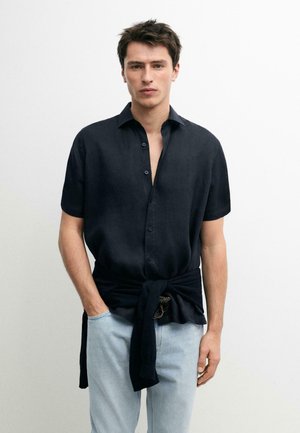 Jeune homme aux cheveux courts et foncés, portant une chemise noire à manches courtes boutonnée et un jean bleu clair, avec un pull noir noué autour de la taille.