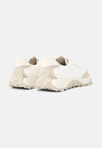 Sneaker bianco con accenti beige, dotato di una suola testurizzata, parte superiore liscia e branding minimale sul tallone. Design con lacci e forma arrotondata.