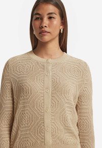 Beige gestrickte Strickjacke mit einem strukturierten hexagonalen Muster, Rundhalsausschnitt und Knopfverschluss an der Vorderseite.