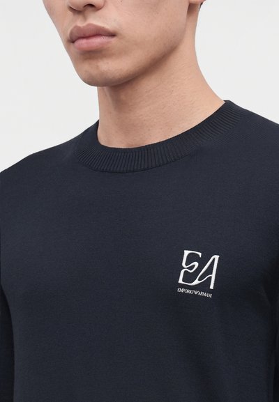 Gros plan d'une personne portant un pull bleu marine foncé avec un col rond côtelé et un logo blanc Emporio Armani EA sur le côté gauche de la poitrine.