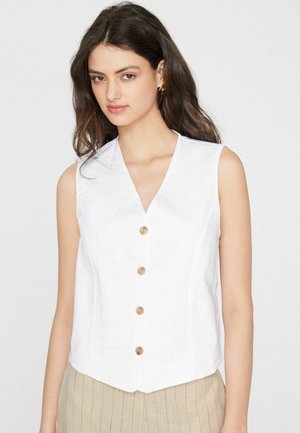 Donna con capelli lunghi e scuri indossa un top bianco senza maniche con bottoni e pantaloni beige a righe, in piedi davanti a uno sfondo chiaro e uniforme.