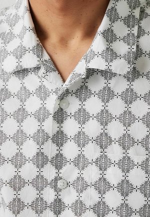 Gros plan d'une personne portant une chemise blanche boutonnée avec un motif complexe de diamants noirs et une poche poitrine.