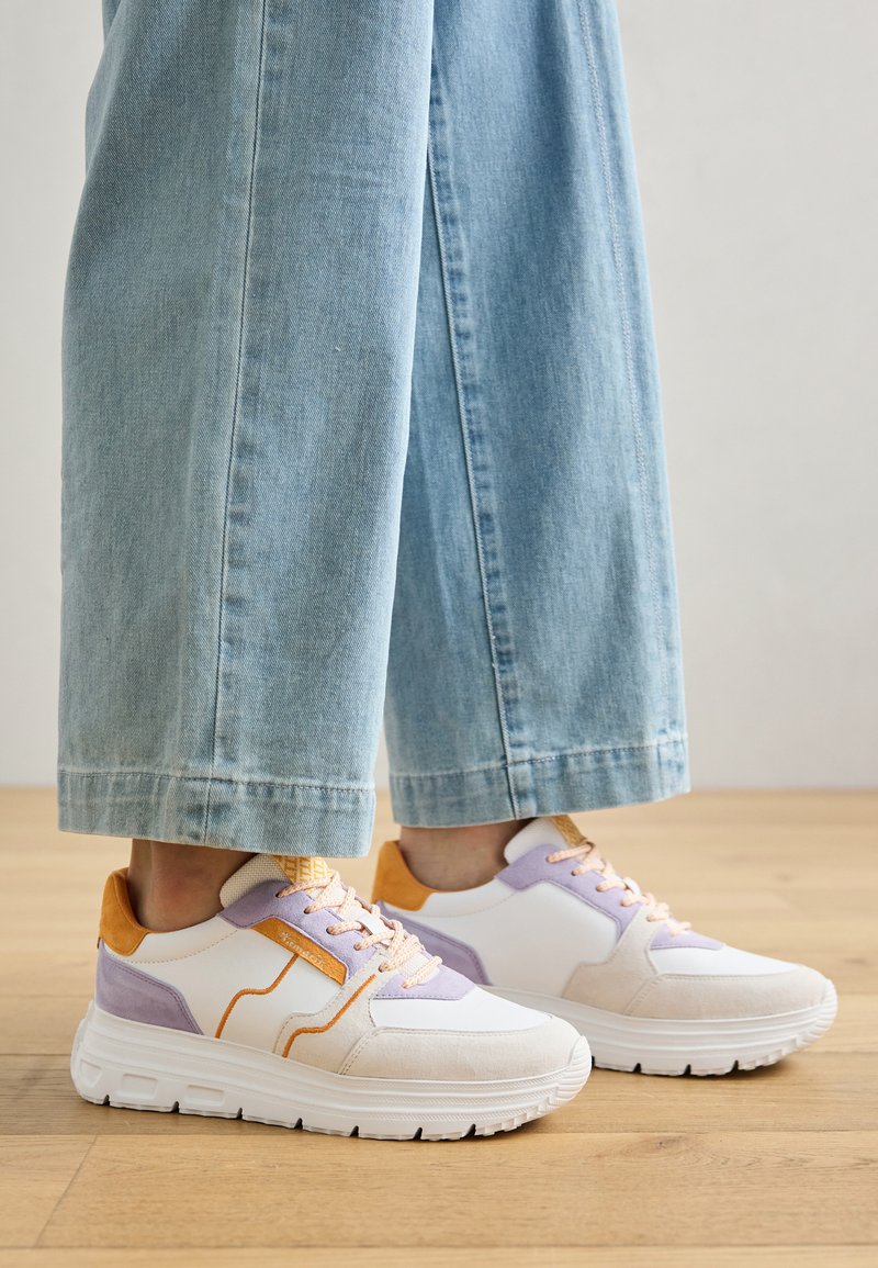 Weiße Sneaker mit lila und orangefarbenen Akzenten, strukturierten Materialien und einer dicken weißen Sohle, kombiniert mit hellblauen weit geschnittenen Jeans.