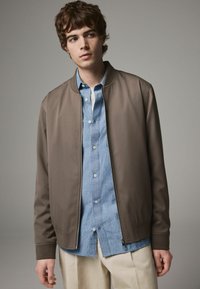 Braune Bomberjacke mit glatter Textur und Reißverschluss. Getragen über einem hellblauen Button-up-Hemd und weißen Hosen.