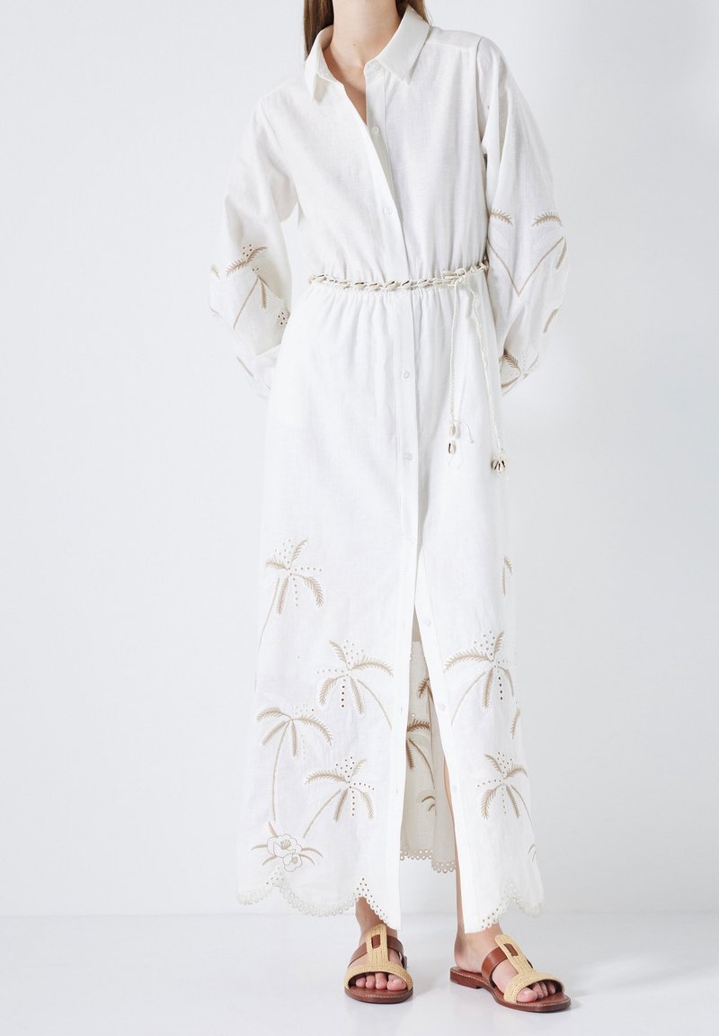 Femme portant une longue robe blanche boutonnée avec des palmiers brodés et une ceinture en corde, associée à des sandales tressées à bouts ouverts.