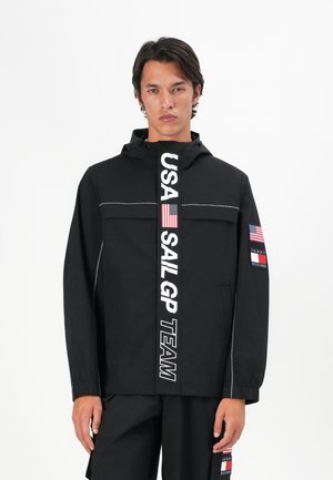 Schwarze Kapuzenjacke mit durchgehendem Reißverschluss, weißen Textmarkierungen und Akzenten der amerikanischen Flagge an den Ärmeln. Glatte Textur, sportliches Design.