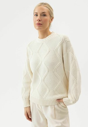 Femme portant un pull en maille torsadée crème avec un col rond et des manches ballon, associée à un pantalon clair, debout devant un fond blanc.