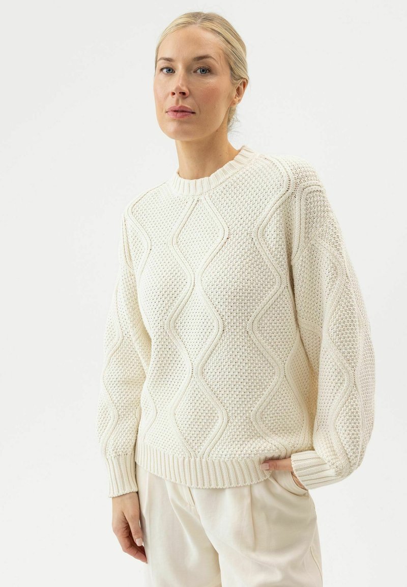 Femme portant un pull en maille torsadée crème avec un col rond et des manches ballon, associée à un pantalon clair, debout devant un fond blanc.