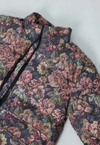 Veste matelassée florale avec un fond noir, ornée de roses multicolores et de feuilles. Détails de fermeture éclair et poche avant inclus.