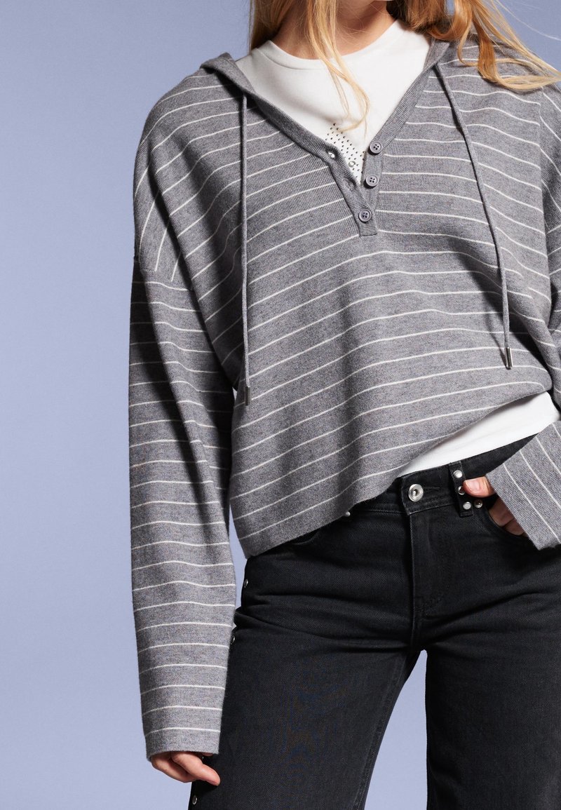 Personne portant un sweat à capuche gris rayé boutonné sur une chemise blanche, associé à un jean noir taille haute, sur un fond clair uni.