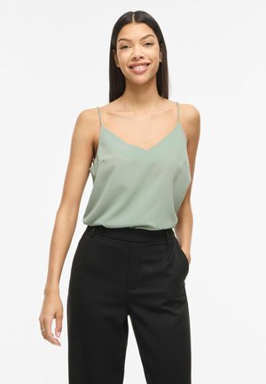 Jeune femme debout souriante, une main dans la poche, portant un haut sans manches vert clair et un pantalon noir taille haute sur fond blanc.