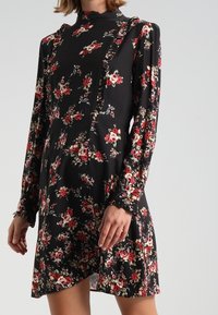 Vestido negro de flores con un cuello alto, mangas largas y detalles con volantes, acentuado con flores rojas y blancas en todo el diseño.