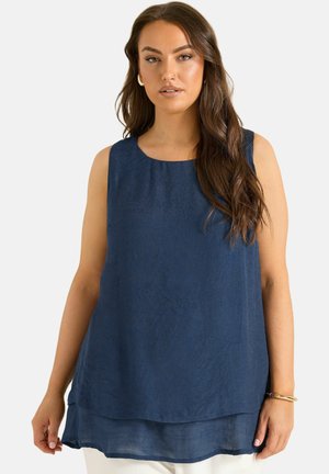 Vrouw met lang bruin haar, draagt een mouwloze marineblauwe gelaagde top en witte broek, staand tegen een effen lichte achtergrond.