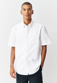 Chemise blanche à manches courtes en tissu de coton, dotée d'une poche poitrine et d'un col classique, associée à un pantalon sombre.
