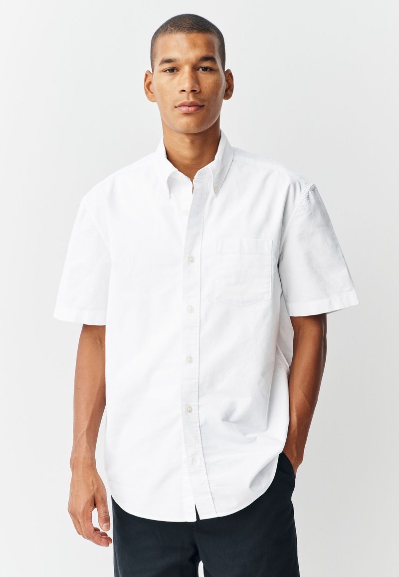 Chemise blanche à manches courtes en tissu de coton, dotée d'une poche poitrine et d'un col classique, associée à un pantalon sombre.