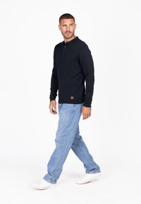 Dunkelblaues Langarm-Polo-Shirt mit drei Knöpfen, kombiniert mit hellblauen Jeans und weißen Sneakern, vor einem weißen Hintergrund stehend.